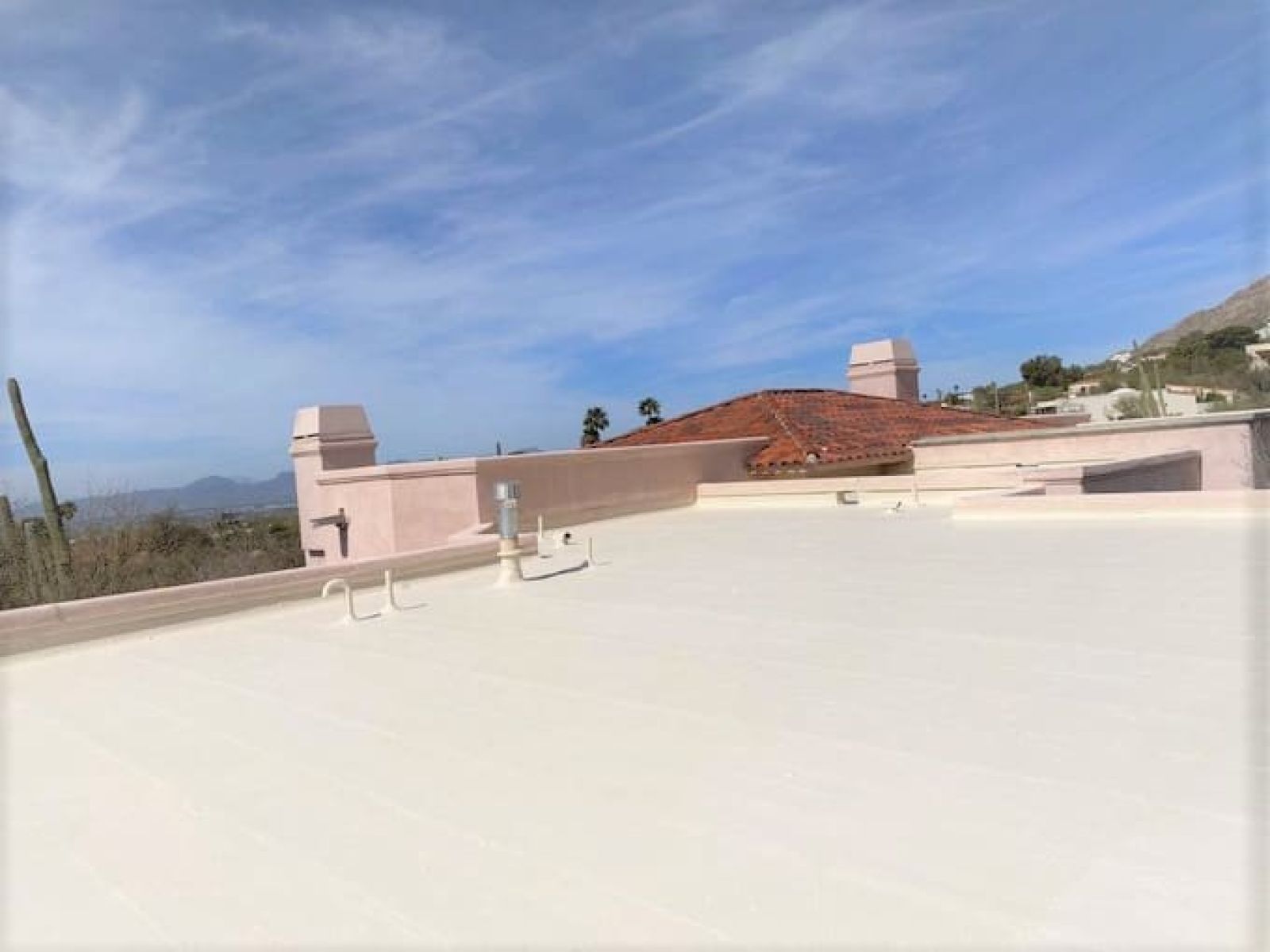 Flat roof with a light-colored surface and various vents, showing a well-maintained roofing surface under a clear sky with a tiled roof and mountains in the background.