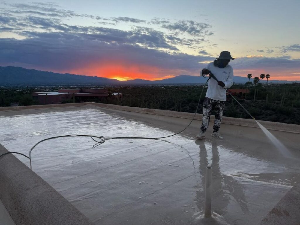 A landscape image of a Tucson home with a flat roof, freshly coated with a reflective or elastomeric roof coating. The setting includes desert landscaping with cacti, a bright sunny sky, and visible heat reflection from the roof.