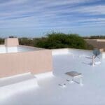 A landscape image of a Tucson home with a roof featuring a freshly applied reflective coating. The scene includes desert landscaping with cacti and succulents, a bright sunny sky, and an air conditioning unit visible near the house, emphasizing energy efficiency.